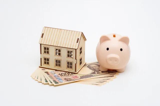 紙幣の上に置かれた家の模型と、横に並ぶピンク色の貯金箱のイメージ