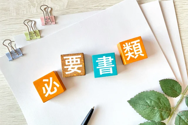 白い紙の上に「必要書類」と書かれた4色の積み木が並んでいる。ペンと書類クリップ、植物の葉が添えられており、事務手続きや準備をイメージさせる。