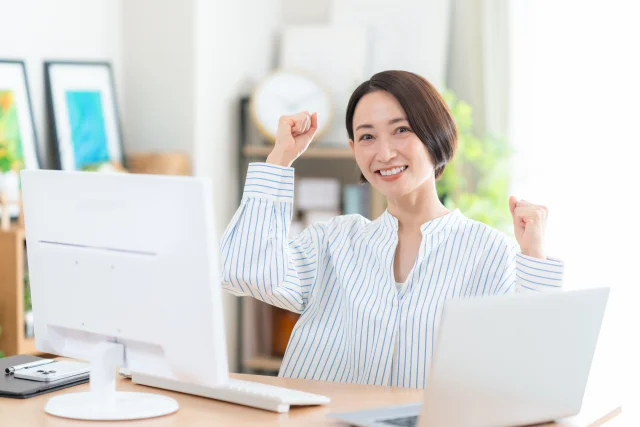 自宅でパソコン作業をする個人事業主の女性が、前向きな表情でガッツポーズしている