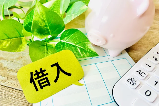 通帳の上に「借入」と書かれた黄色い吹き出しが置かれ、横にピンクの豚の貯金箱、計算機、観葉植物が並んでいる