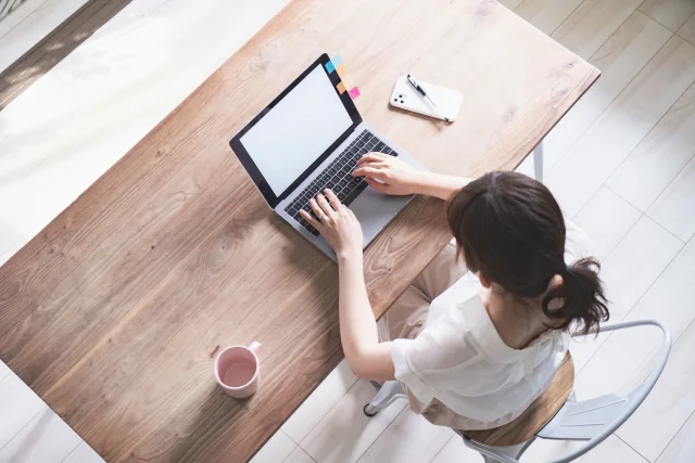 テーブルでノートパソコンを操作する女性の俯瞰写真で、自宅兼事務所の作業風景を表している