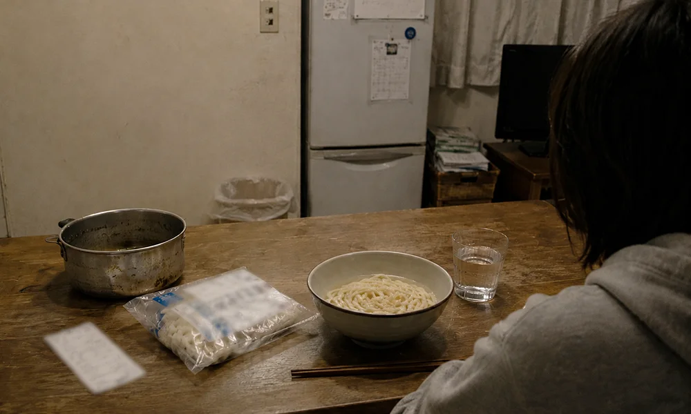 夜の部屋で、具の少ないうどんを前に静かに座る人物の背中。テーブルには鍋、冷凍うどんの袋、レシートが置かれている。