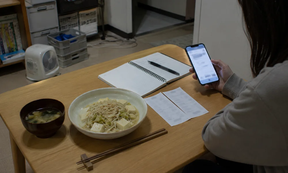 もやしや豆腐を使った質素な食事のそばで、スマホの明細画面と家計メモを確認する手元。